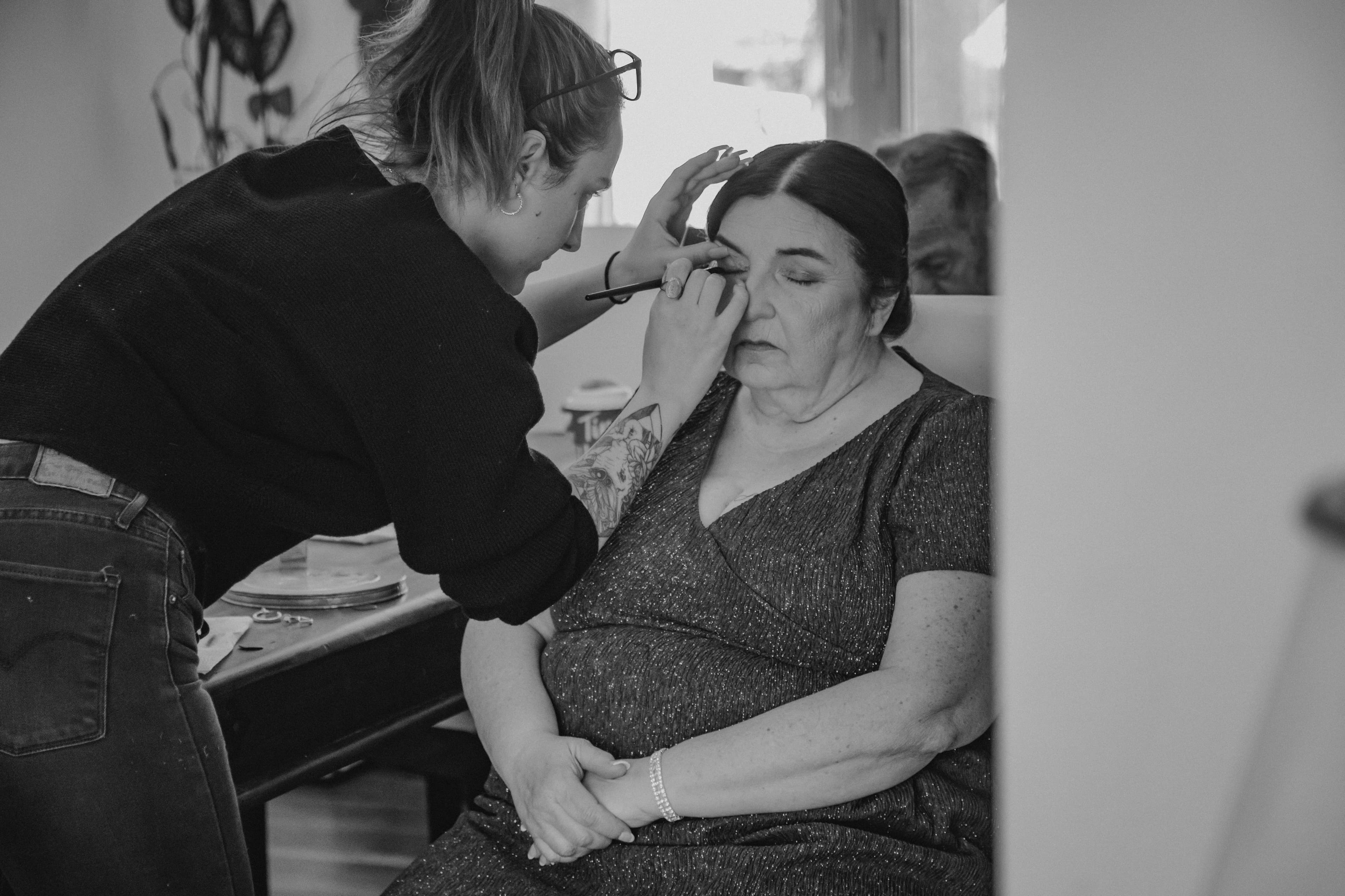 Sia applying eye makeup to a seated client before the ceremony, candid black and white